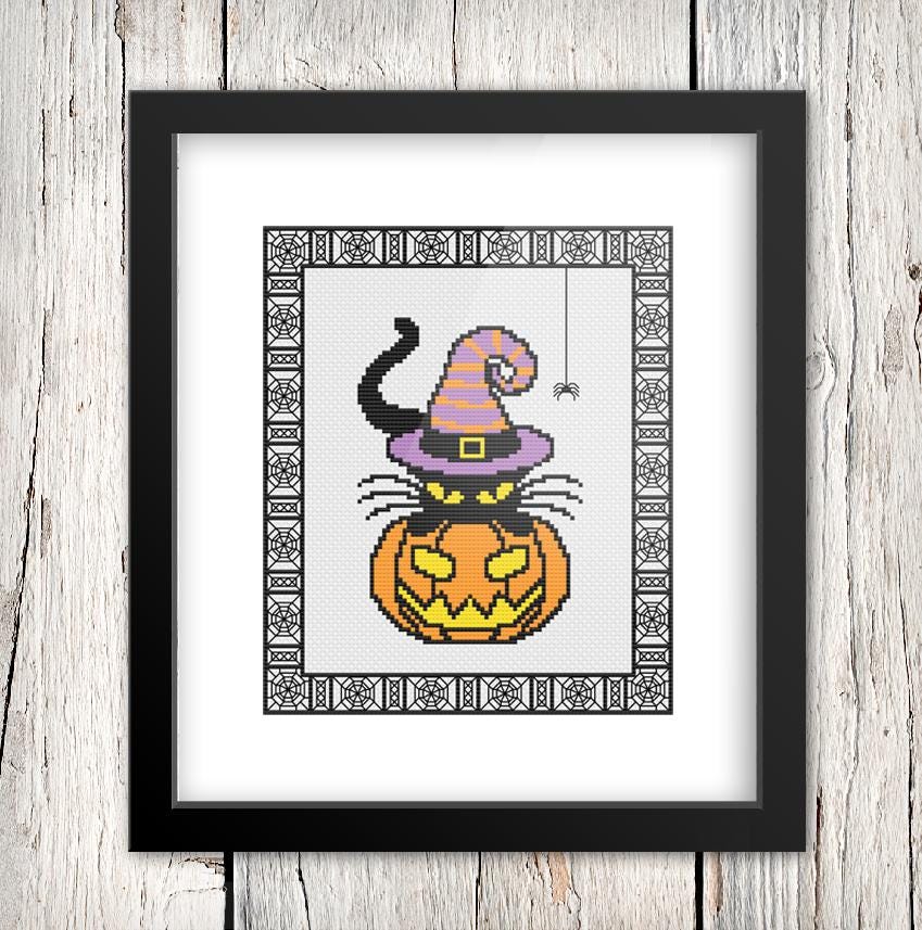 A framed cross-stitch image depicting a witch's hat on top of a jack-o-lantern, with a spider web design around the edges.