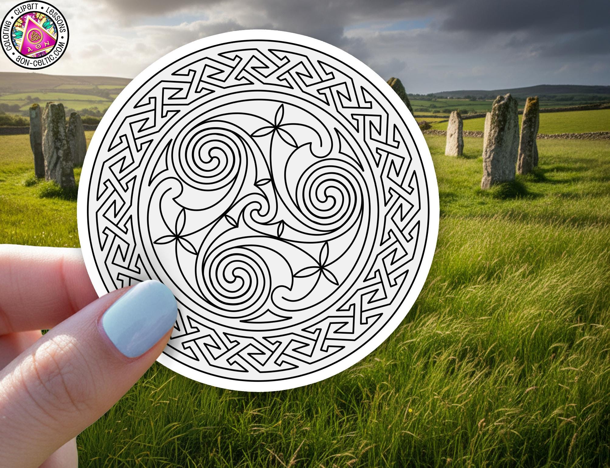 A hand holding a circular design with intricate swirls and floral patterns, set against a backdrop of a grassy field with stone markers in the distance.