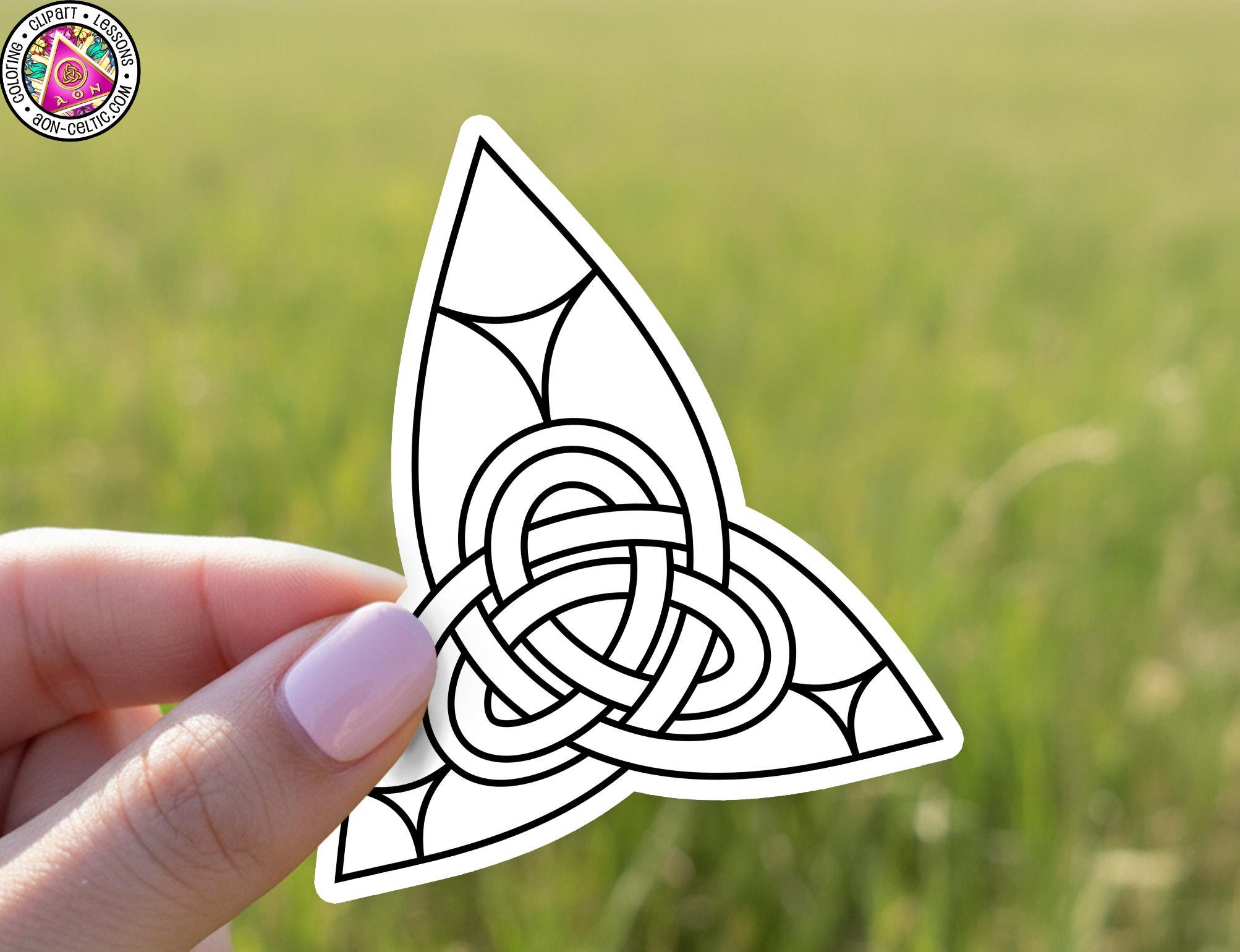 A hand holding a sticker with a Celtic knot design against a blurred green field background.