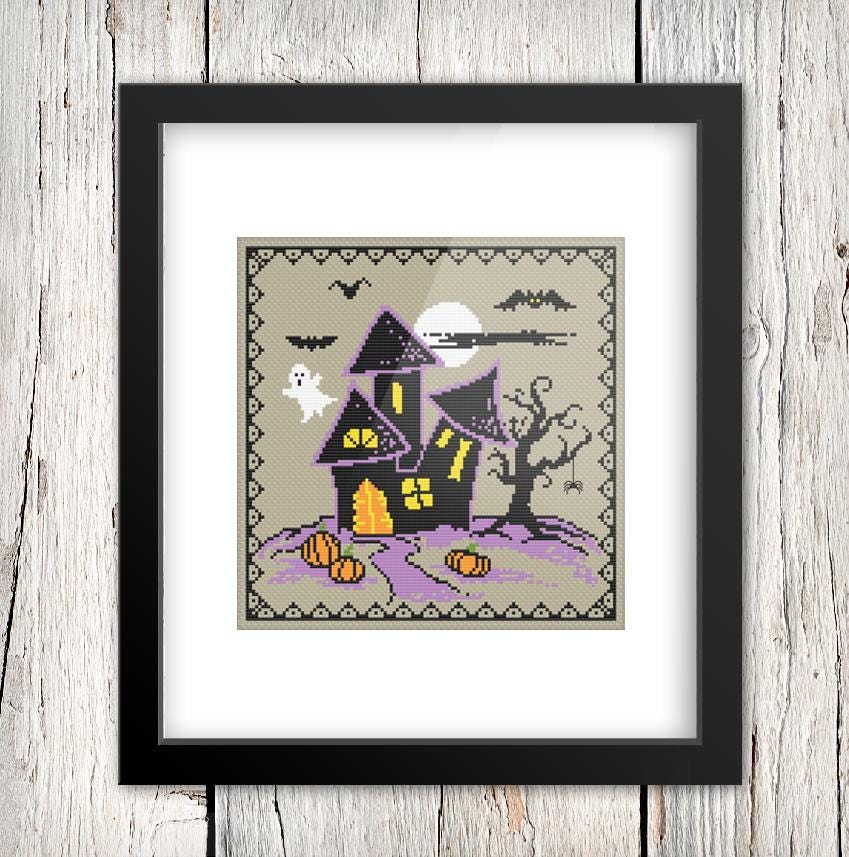 The image is a framed cross-stitch depicting a spooky Halloween scene with a house, bats, a ghost, and pumpkins.
