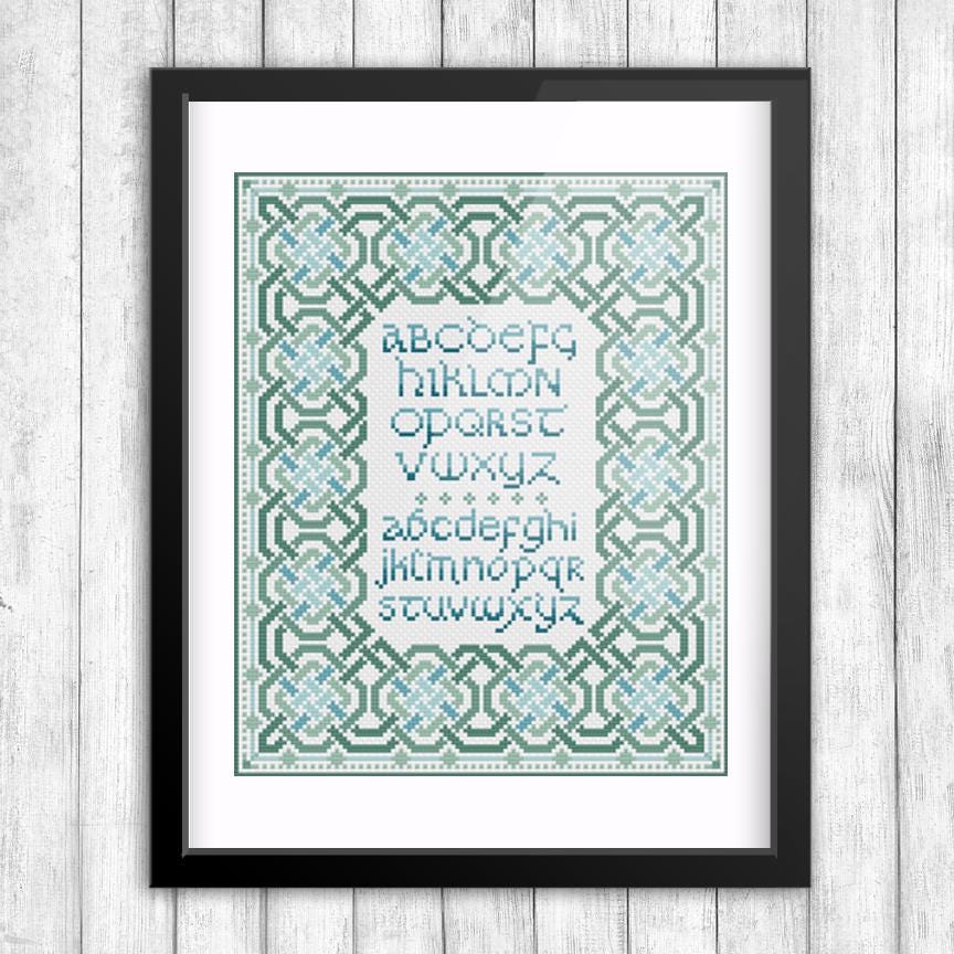 The image is a framed cross-stitch design featuring the alphabet in a decorative pattern.