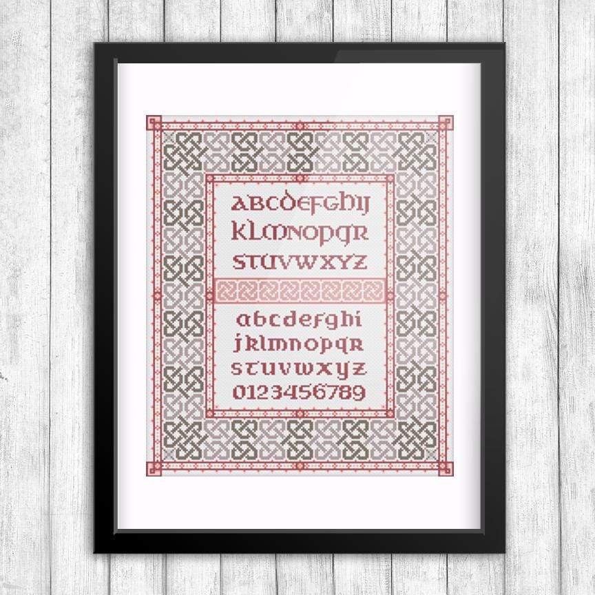 The image is a framed cross-stitch design featuring the alphabet in a decorative pattern.