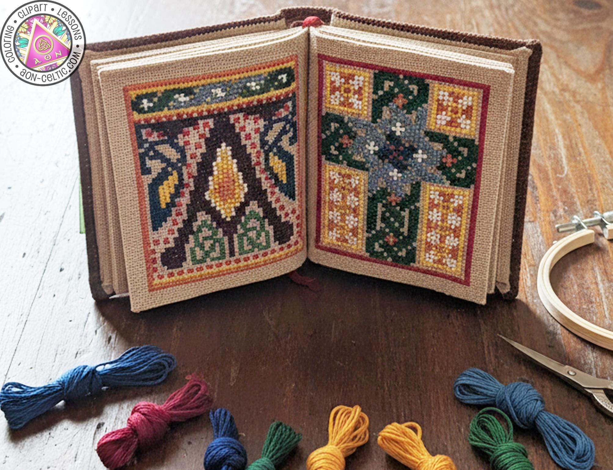 a cross-stitch book with two pages displaying intricate patterns, along with various embroidery floss and a pair of scissors on a wooden surface.