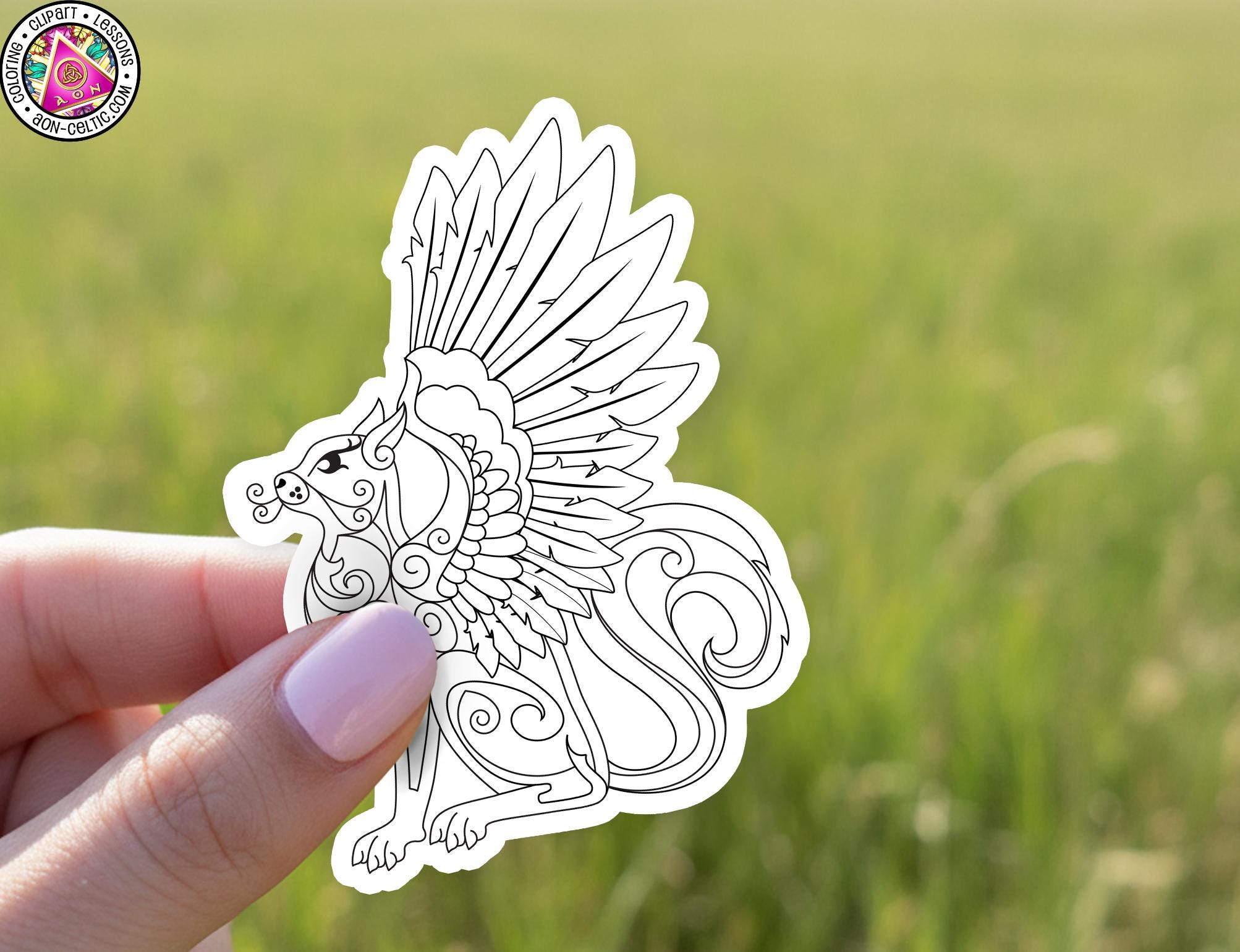 A hand holding a white sticker with a detailed black and white illustration of a winged creature against a blurred green background.