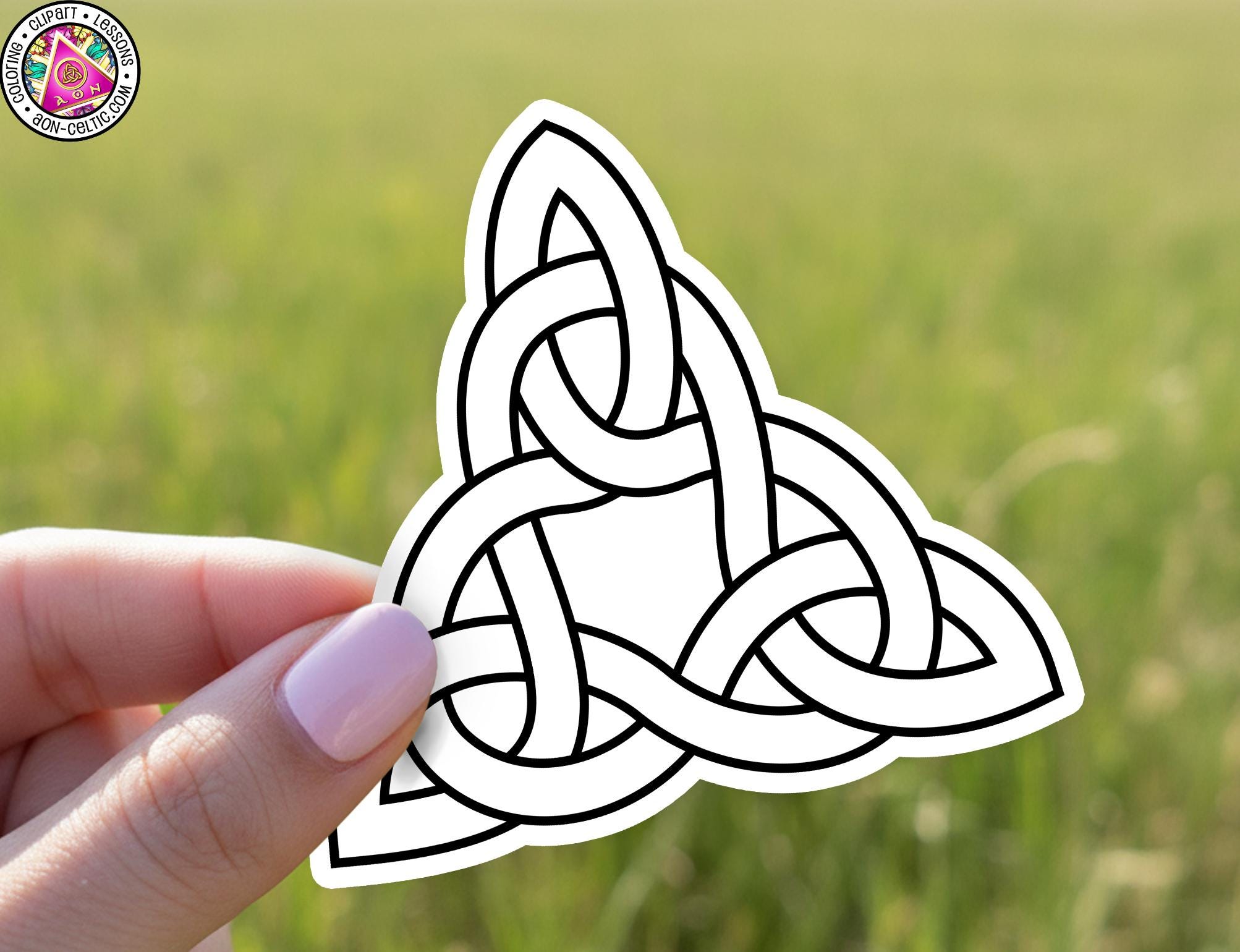 A hand holding a sticker with a black and white design of a Celtic knot against a blurred green background.
