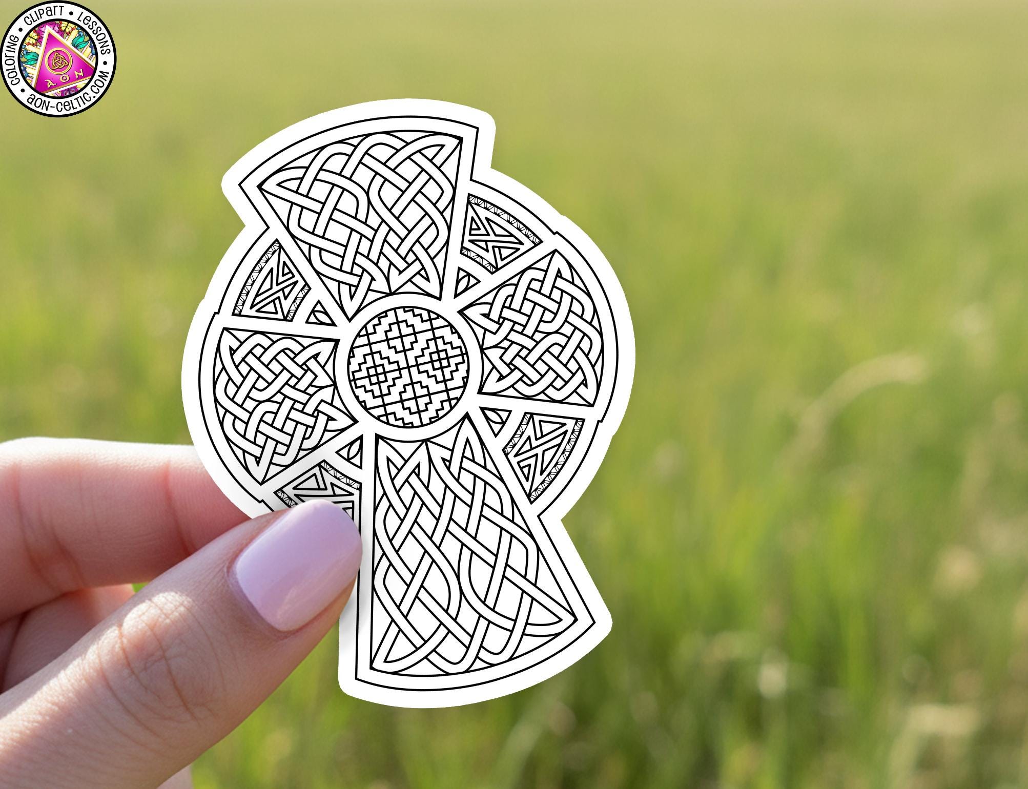 A hand holding a sticker with a detailed Celtic knot design against a blurred green field background.
