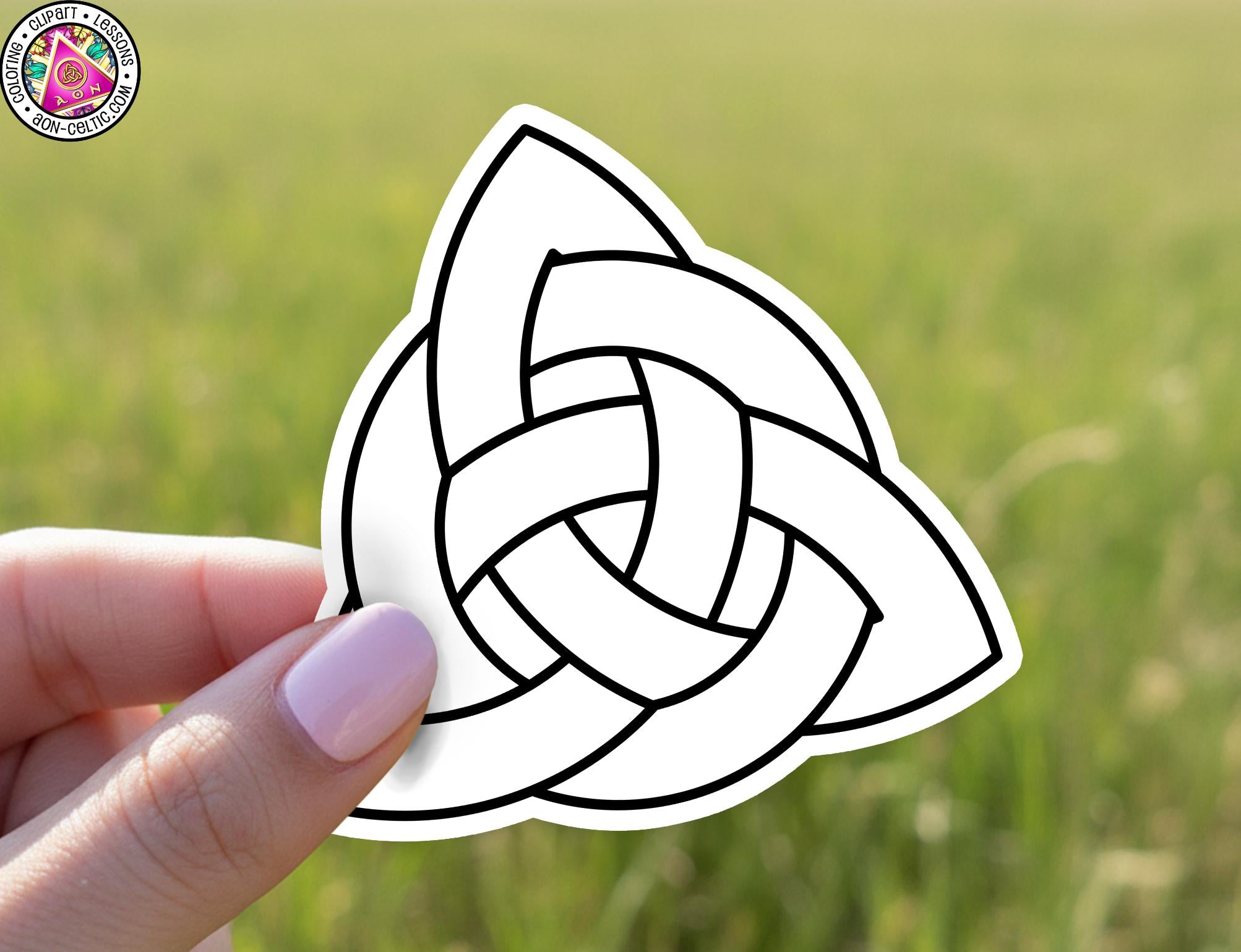 A hand holding a sticker of a Celtic knot against a blurred green field background.