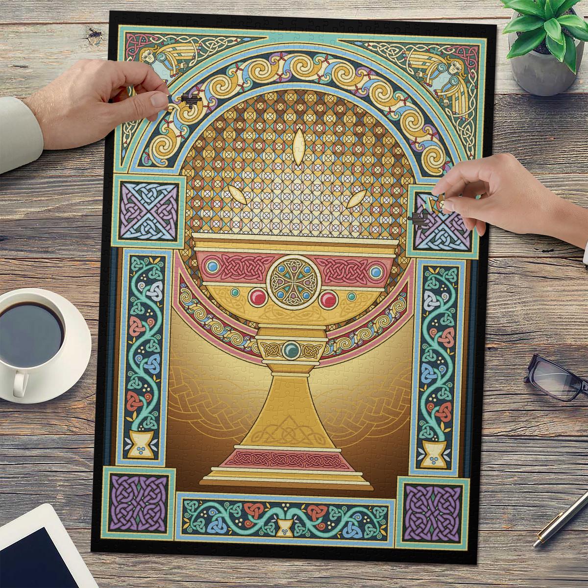 a large, ornate, and colorful illustration of a golden bowl or vase, surrounded by intricate patterns and designs. The artwork is displayed on a wooden surface, with a pair of hands reaching towards it. There are also various objects in the background, including a cup, a plant, and a pair of glasses.