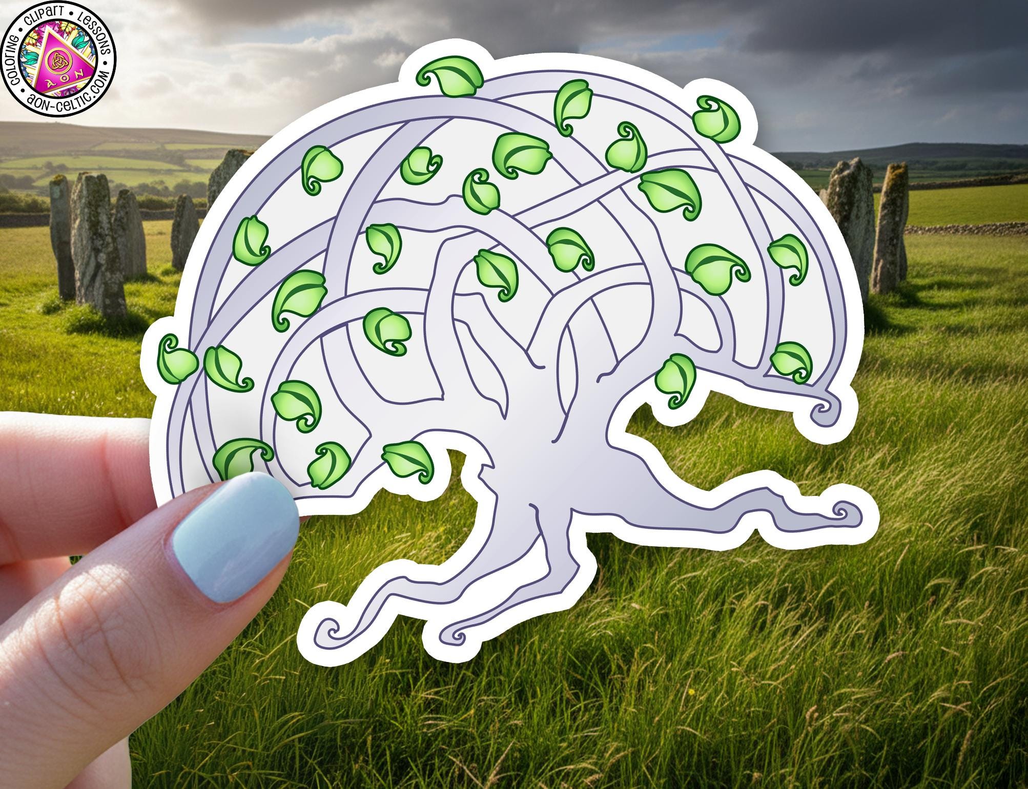 A hand holding a white sticker with a stylized tree design, surrounded by green leaves, against a backdrop of a grassy field with stone markers.