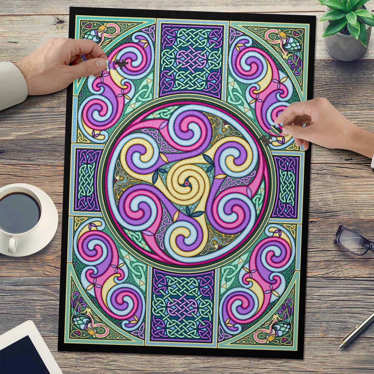 a colorful, intricate Celtic-inspired design with swirling patterns and shapes in shades of pink, purple, green, and yellow. The design is displayed on a large poster or canvas, and two hands are visible, one on the left and one on the right, suggesting that someone is examining or working on the design.