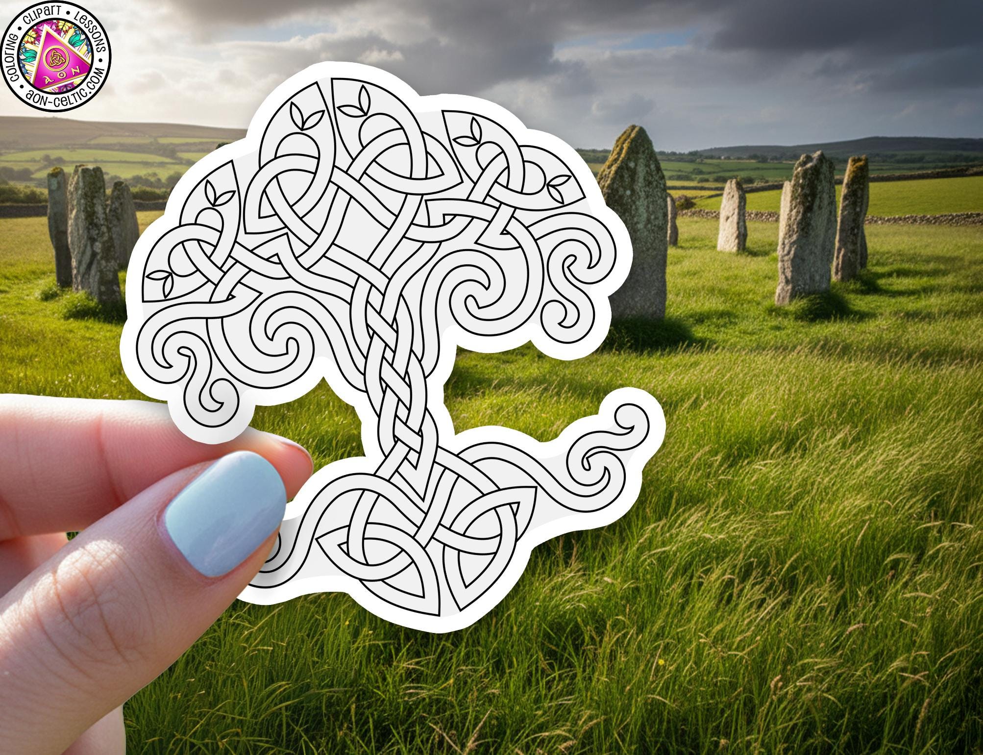 A hand holding a white paper with a black and white tree design, with a grassy field and stone markers in the background.