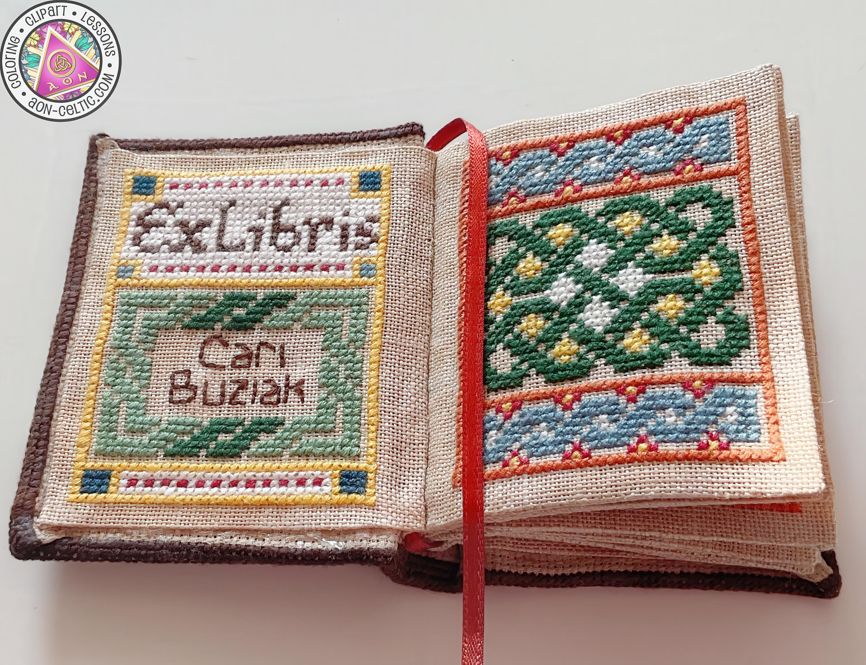 a hand-stitched book with a cross-stitch design, featuring a red ribbon bookmark.