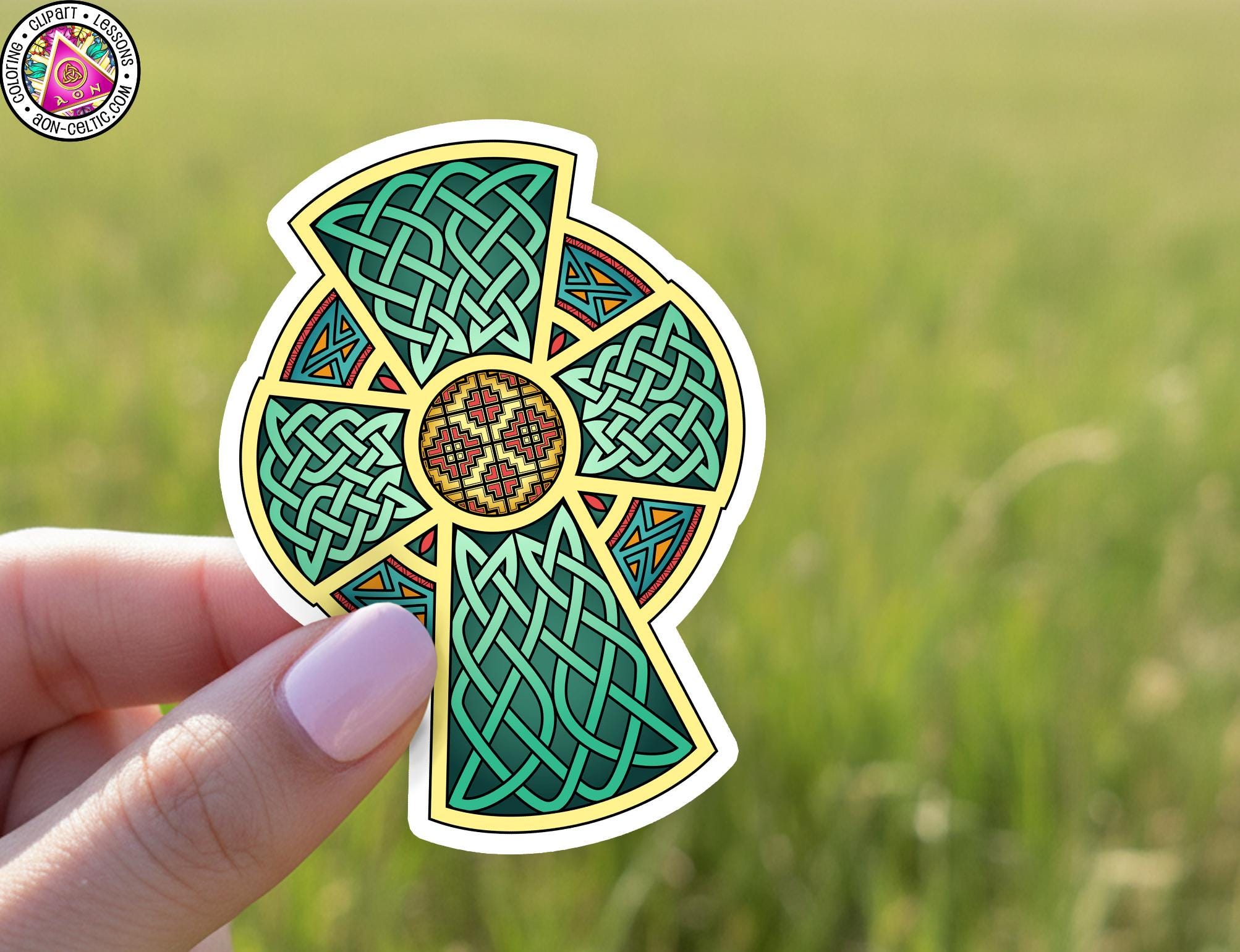 A hand holding a colorful sticker with a Celtic cross design against a blurred green field background.