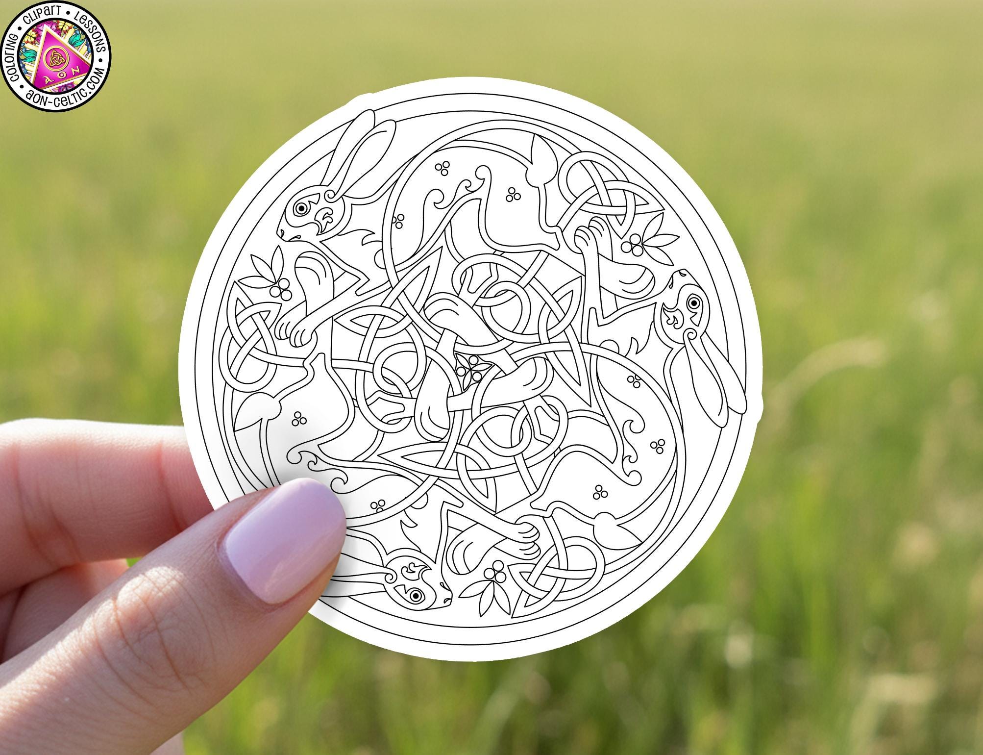 A hand holding a circular design featuring intricate Celtic knot patterns against a blurred green field background.
