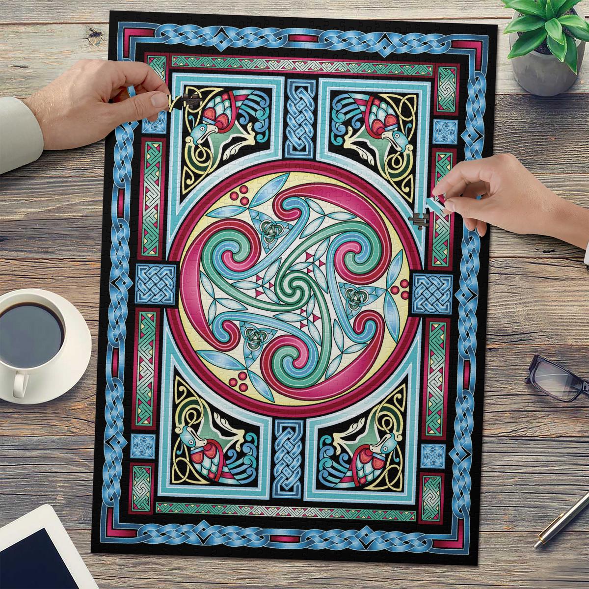 a colorful, intricate Celtic-inspired design with swirling patterns and shapes in shades of blue, pink, and green. The design is displayed on a large, framed piece of paper, and two hands are reaching towards it, suggesting someone is examining or working on the artwork.