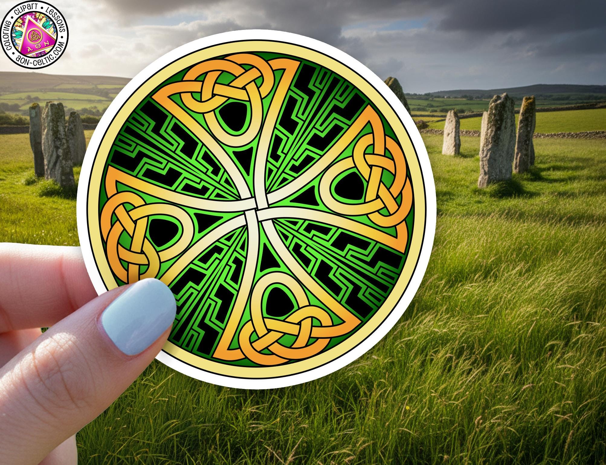 A hand holding a circular sticker with a colorful, intricate Celtic knot design against a backdrop of a grassy field with stone markers.
