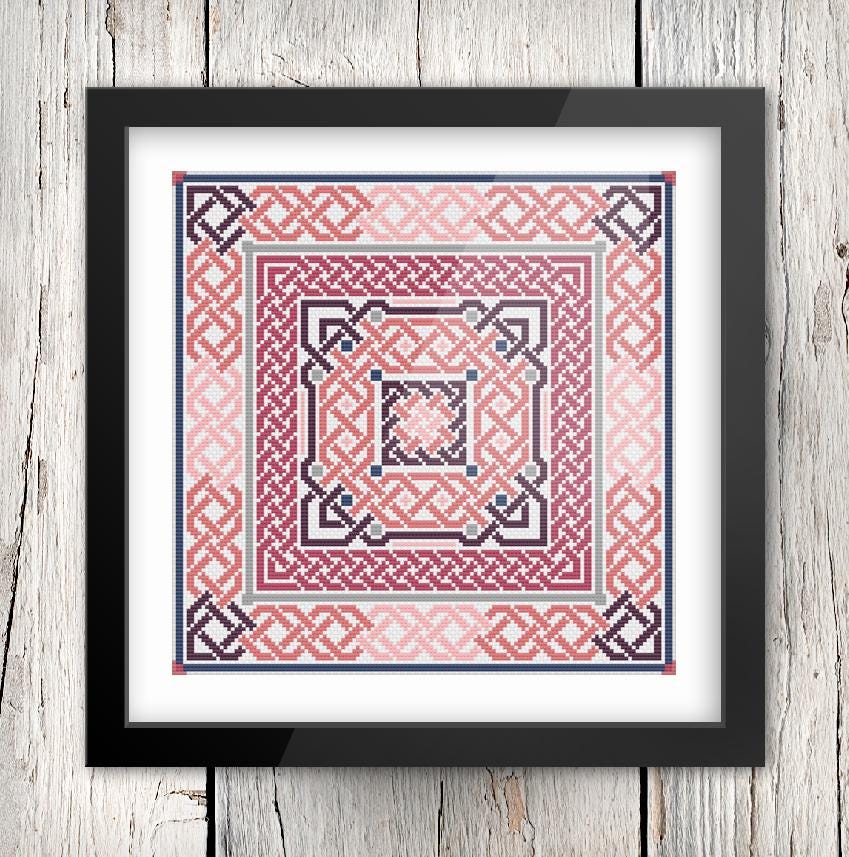 The image is a framed cross-stitch design featuring a repeating pattern of geometric shapes in shades of pink, purple, and blue.