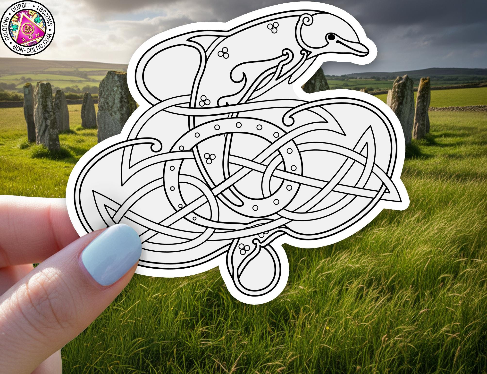 A hand holding a sticker with a stylized illustration of a dolphin or sea creature, surrounded by intricate Celtic knot patterns, against a backdrop of a grassy field with stone markers.