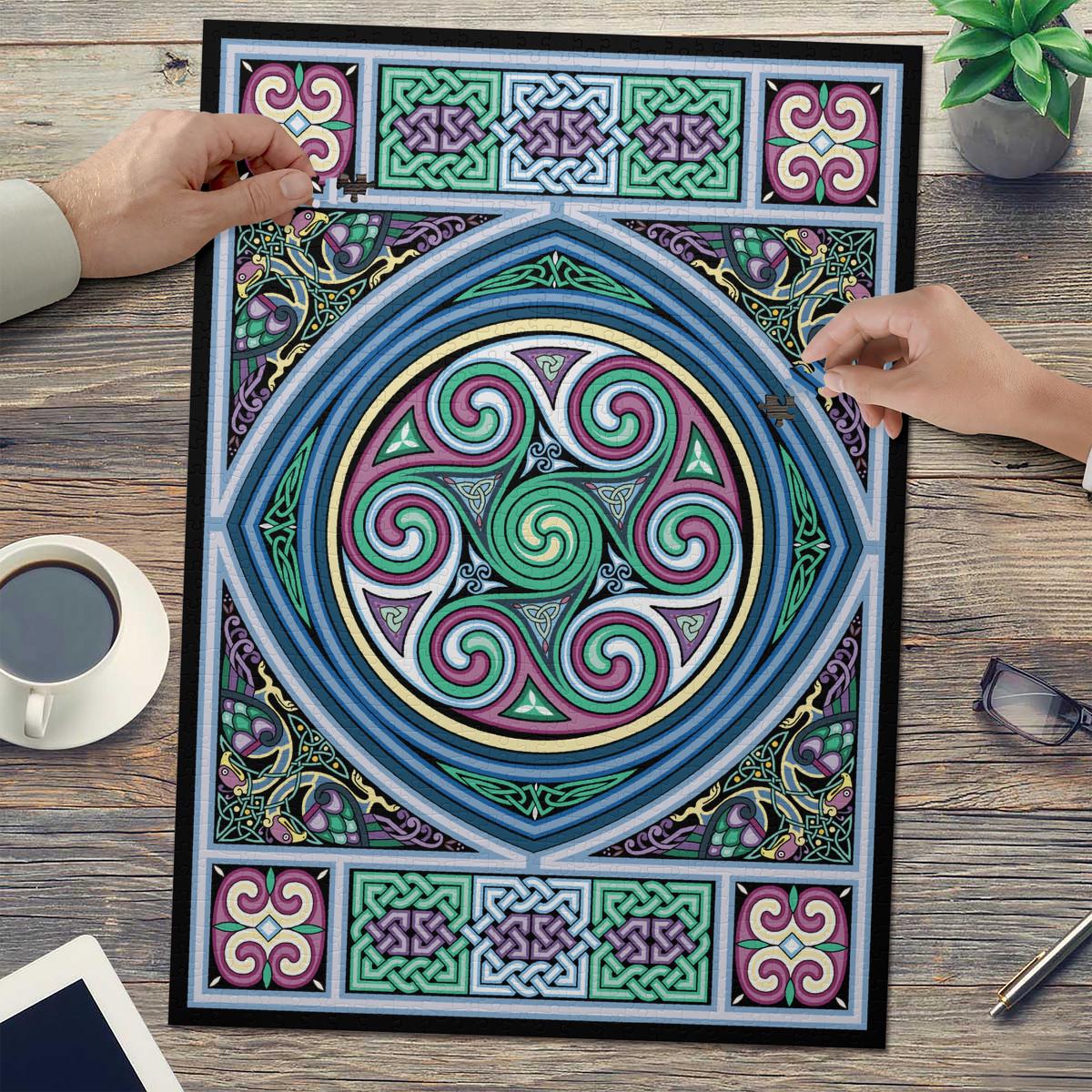 a large, colorful, and intricate Celtic-inspired design with swirling patterns and shapes in shades of blue, green, purple, and pink. The design is displayed on a poster or canvas, and two hands are visible, one on the left and one on the right, suggesting that someone is working on or admiring the design.