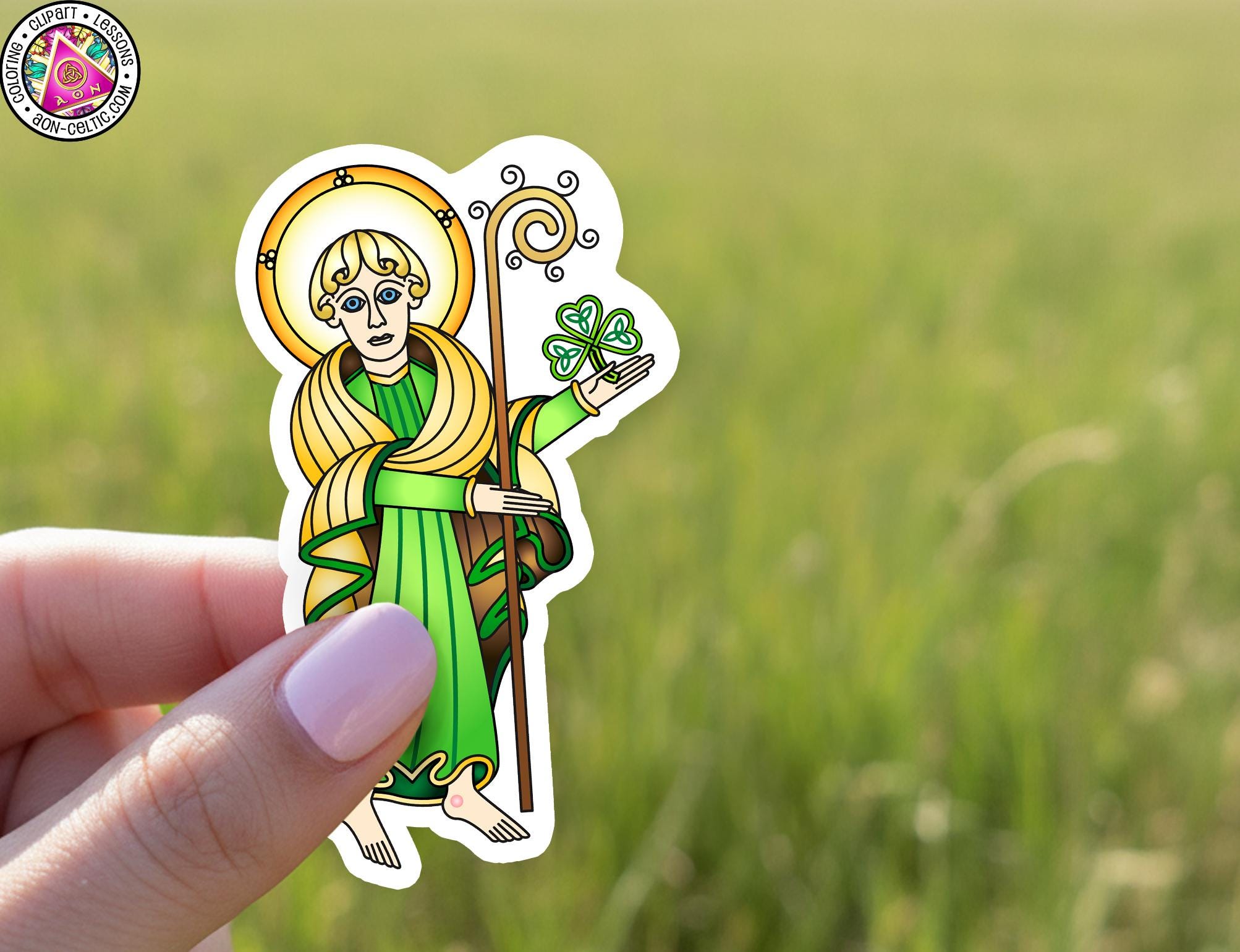 A hand holding a sticker of a saint with a clover leaf in their hand, set against a blurred green background.