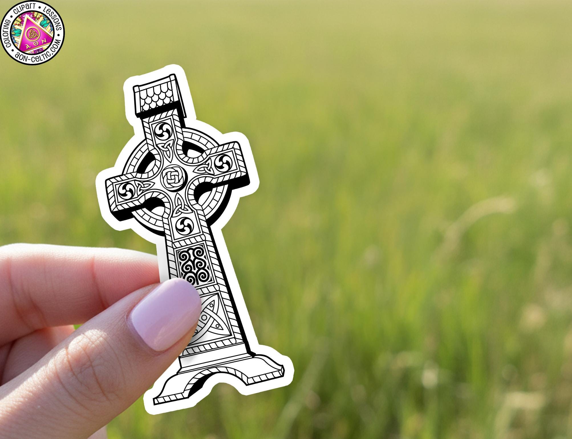 A hand holding a sticker of a Celtic cross against a blurred green field background.