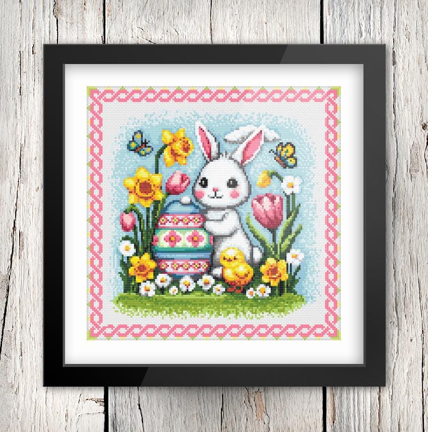 A framed cross-stitch image depicting a white rabbit sitting on a grassy area surrounded by flowers, including yellow daffodils, pink tulips, and yellow chicks.