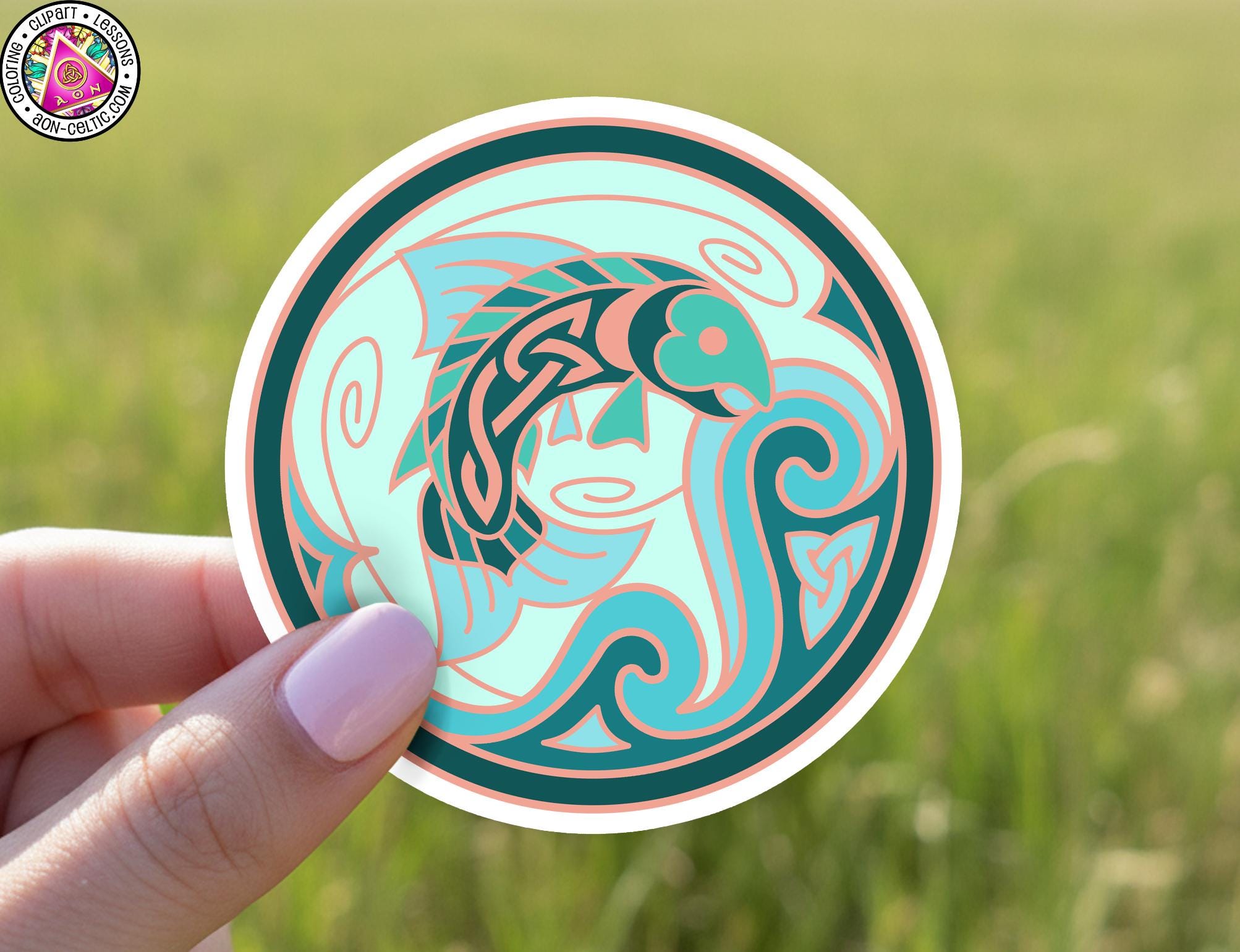 A hand holding a circular sticker with a fish design on it, set against a blurred green field background.