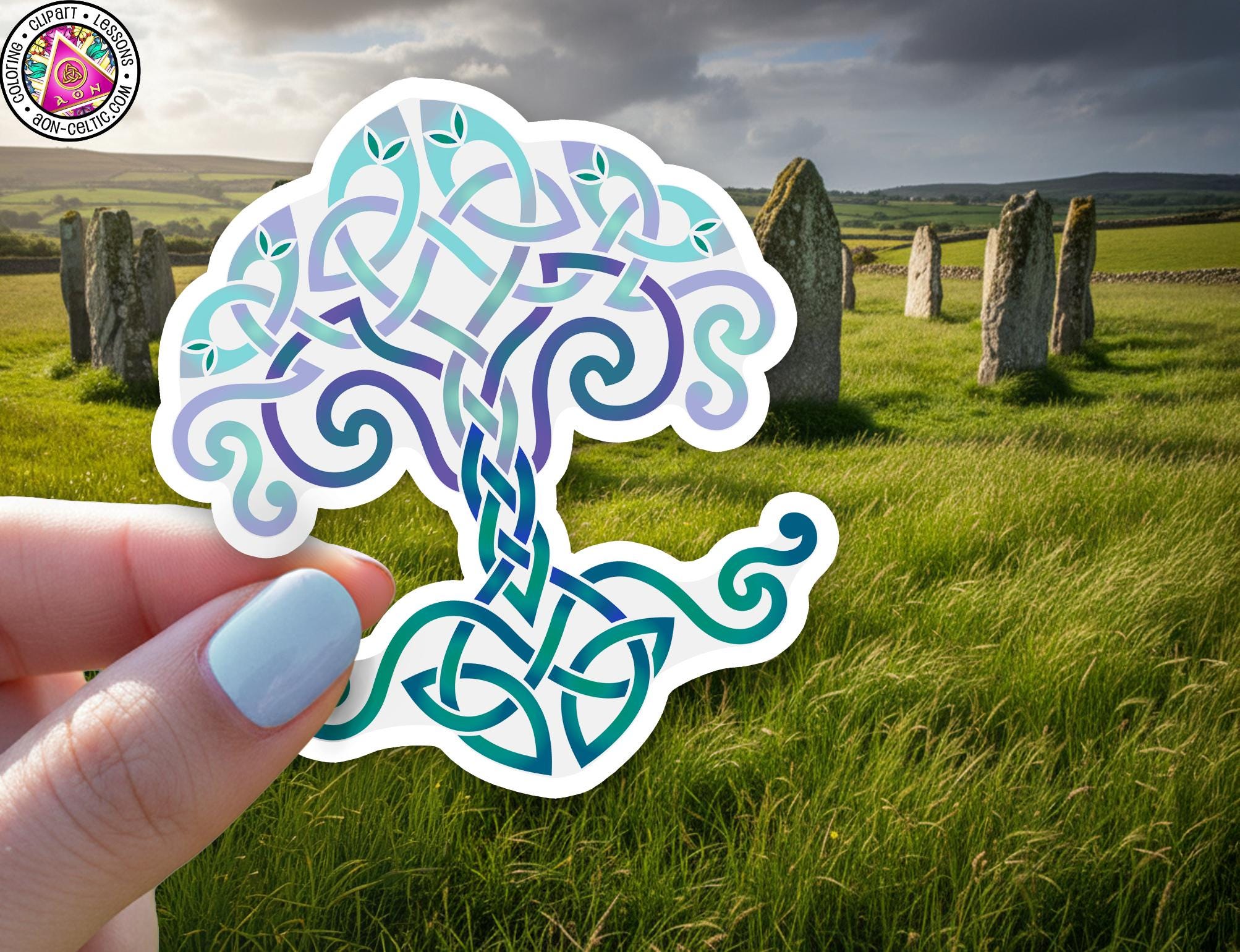 A hand holding a sticker with a Celtic tree design against a backdrop of a grassy field with stone markers in the distance.