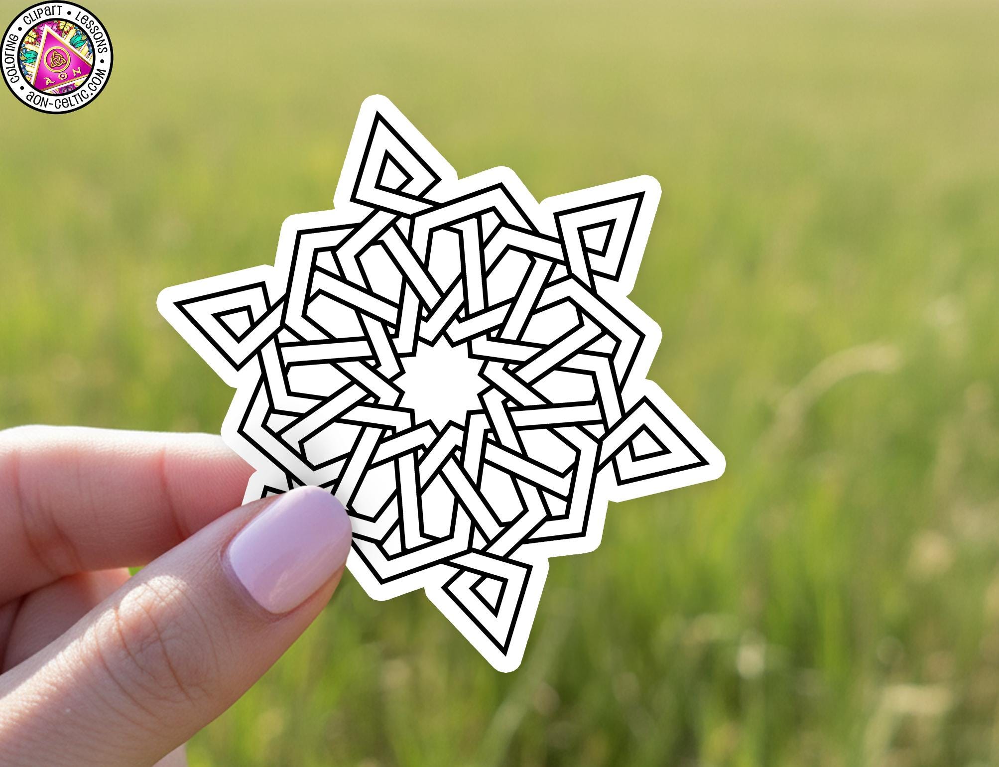 A hand holding a white sticker with a black geometric design against a blurred green field background.
