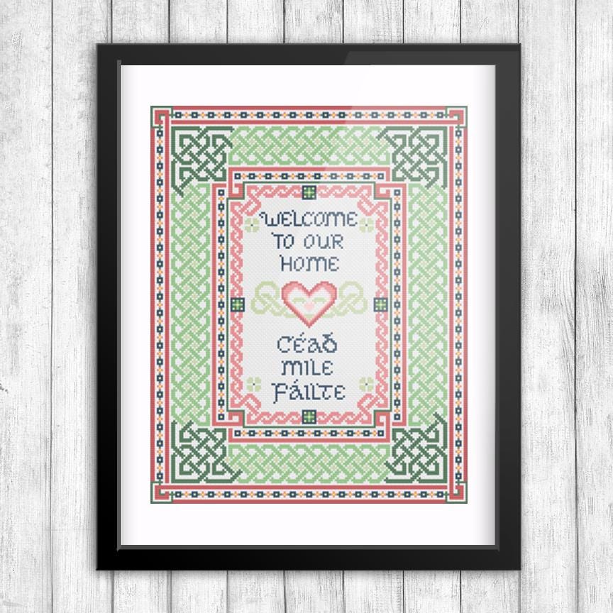 The image is a framed cross-stitch design featuring a heart-shaped motif surrounded by intricate patterns in shades of green and red. The text within the design reads &quot;Welcome to our Home&quot; and &quot;Céad méle fáilte&quot;, which translates to &quot;Welcome to our home&quot; and &quot;Home with love&quot;.