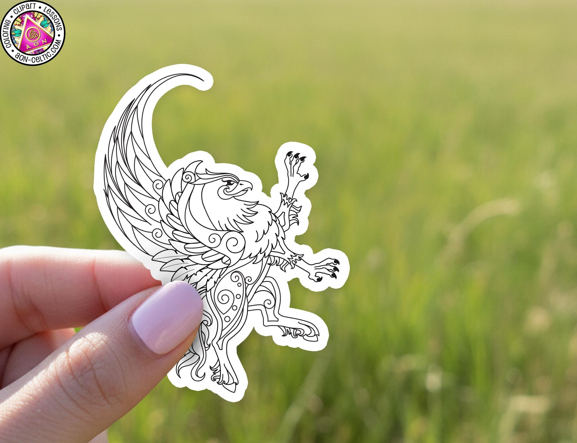 A hand holding a sticker of a winged creature against a blurred green background.