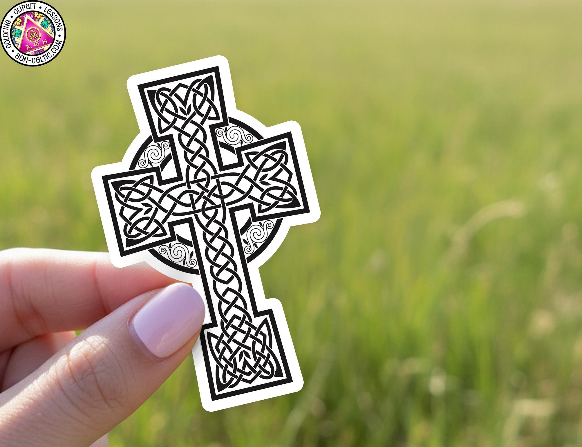 A hand holding a sticker of a Celtic cross against a blurred green field background.
