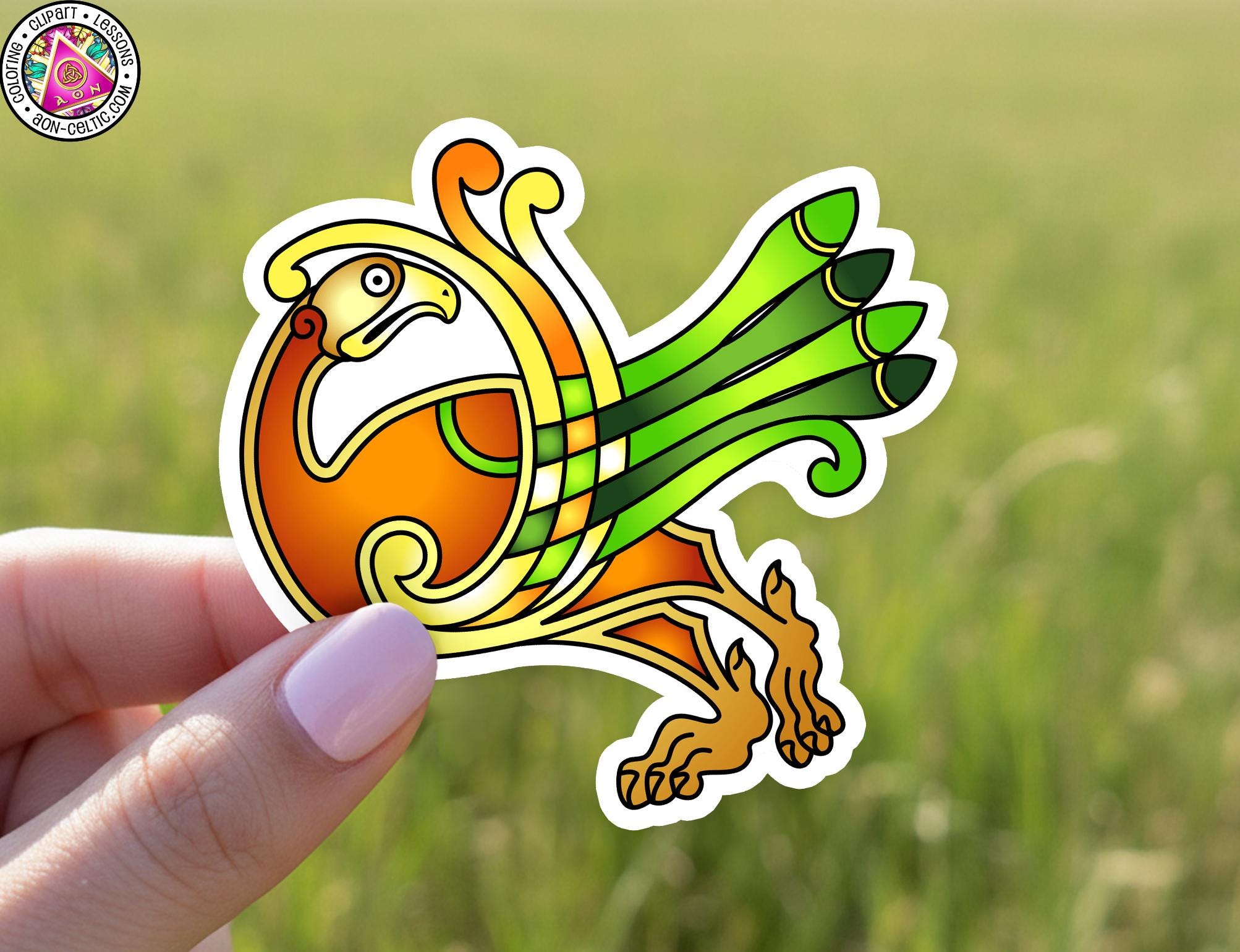 A hand holding a colorful sticker depicting a stylized bird or mythical creature with a yellow body, green wings, and orange accents.