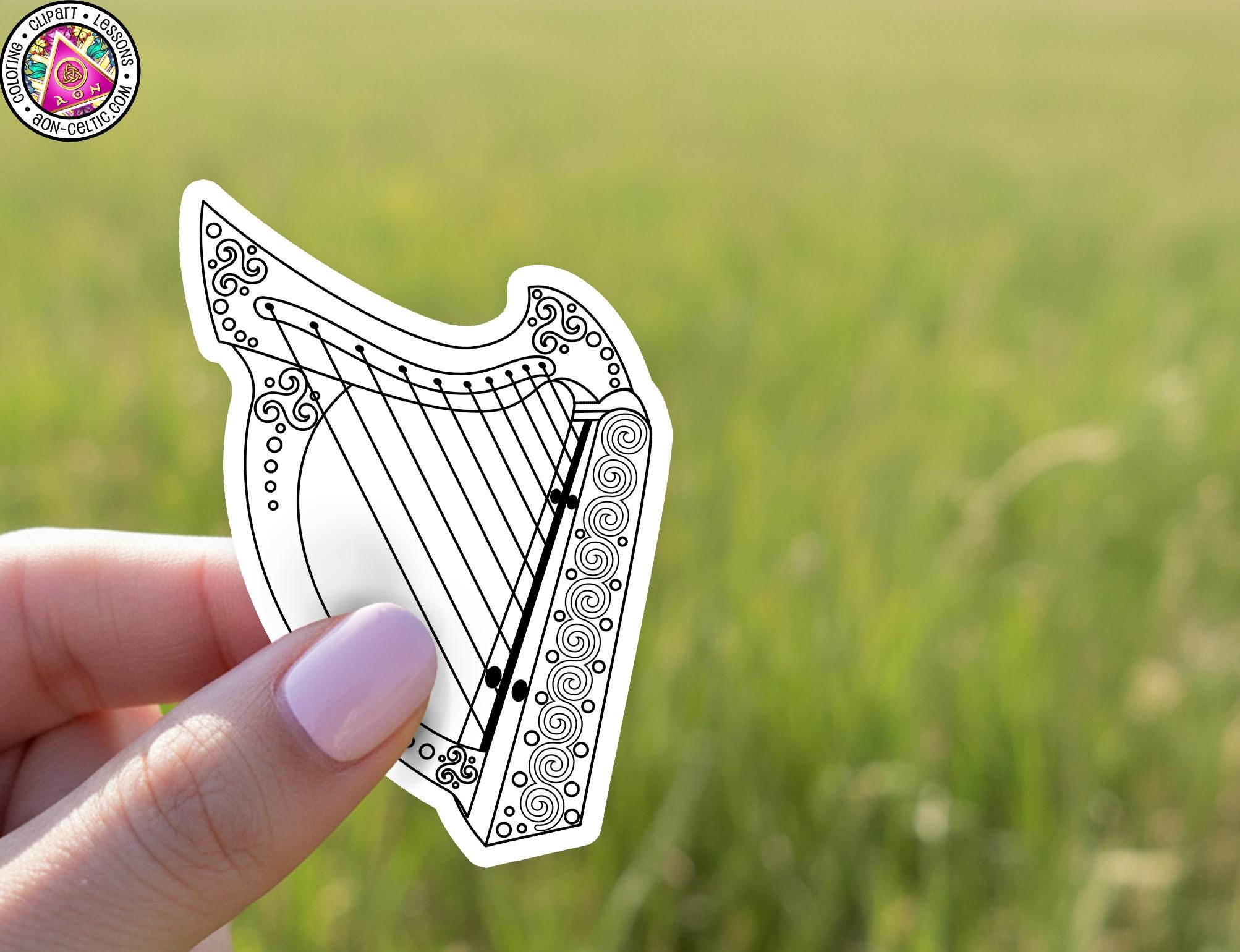A hand holding a sticker of a harp against a blurred green field background.